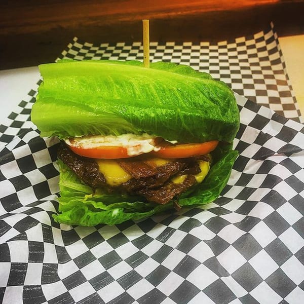 A lettuce-wrapped Burger Bunker burger with cheese, bacon, tomato, and sauce on checkered paper.